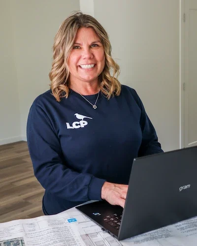 A woman with wavy blonde hair, wearing a navy blue LCP sweatshirt, smiles while working on a laptop at a table with papers in a bright room.