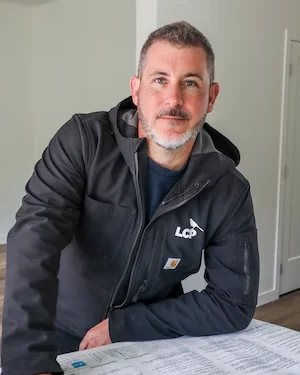 A man with short gray hair and a beard, wearing a dark jacket with an LCP logo, leans on a table covered with papers in a bright, modern room.
