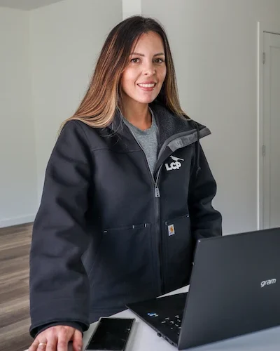 A woman with long brown hair, wearing a black jacket, stands indoors next to a laptop and a smartphone on a white table, smiling at the camera. The room has light walls and wood flooring.