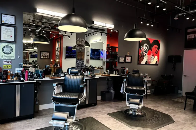 Modern barbershop interior with two black barber chairs, large mirrors, overhead lights, and various grooming products on counters. Framed art and certificates decorate the walls; reflections show more of the stylish space.
