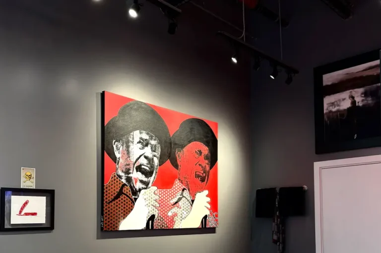 A gallery wall with a large painting of two men in hats on a red background, both shown singing or shouting. Smaller framed artworks are visible nearby under track lighting in a dark-walled room.