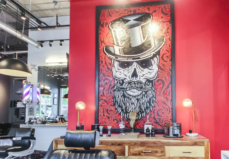 A barbershop interior with modern decor, featuring a large artwork of a skull with a beard and top hat on a red wall, wooden workstations, barber chairs, and vintage-style lamps.