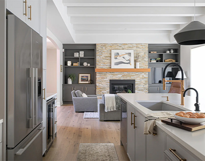 Modern kitchen and living area with gray cabinets, stainless steel appliances, a stone fireplace, wooden floors, and a light gray sofa. Sunlight fills the space, highlighting clean lines and minimalist decor.