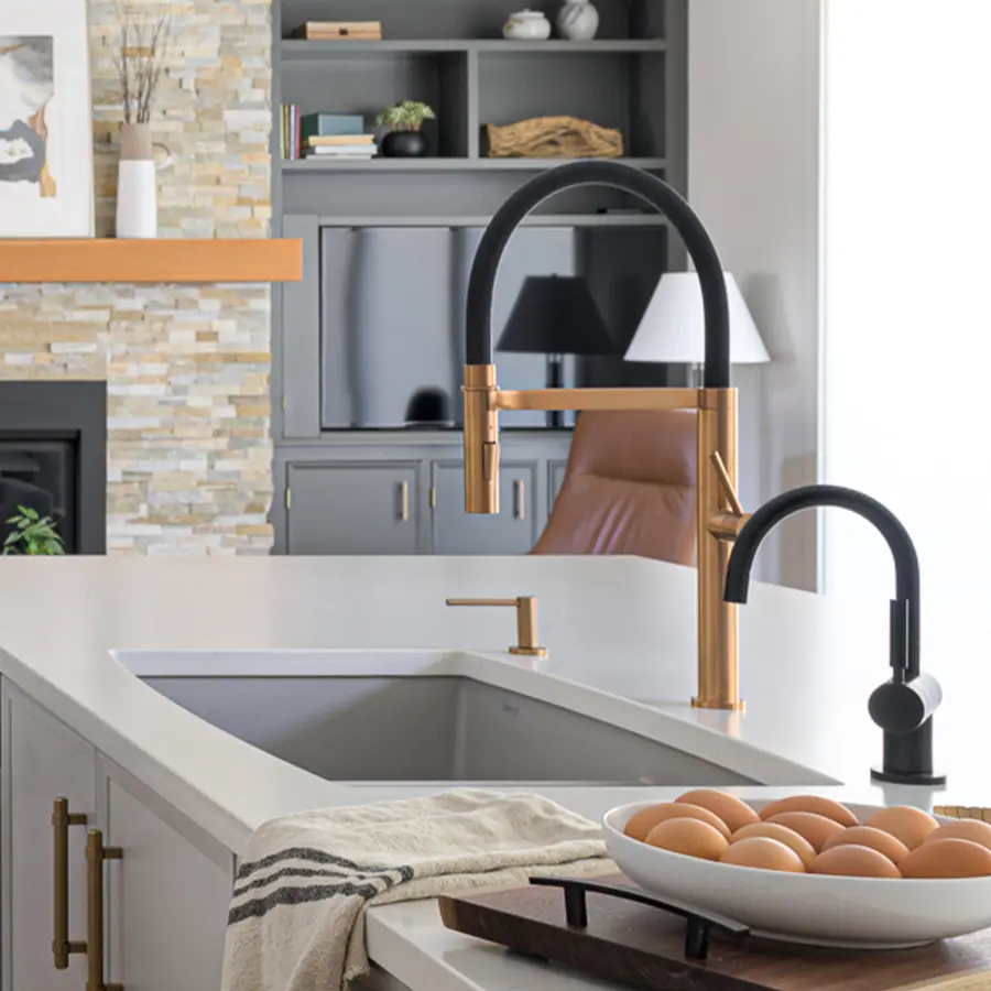 A modern kitchen with a white countertop, gold and black faucet, a striped towel, and a bowl of brown eggs. In the background, gray cabinets, shelves, and a stone fireplace are visible.