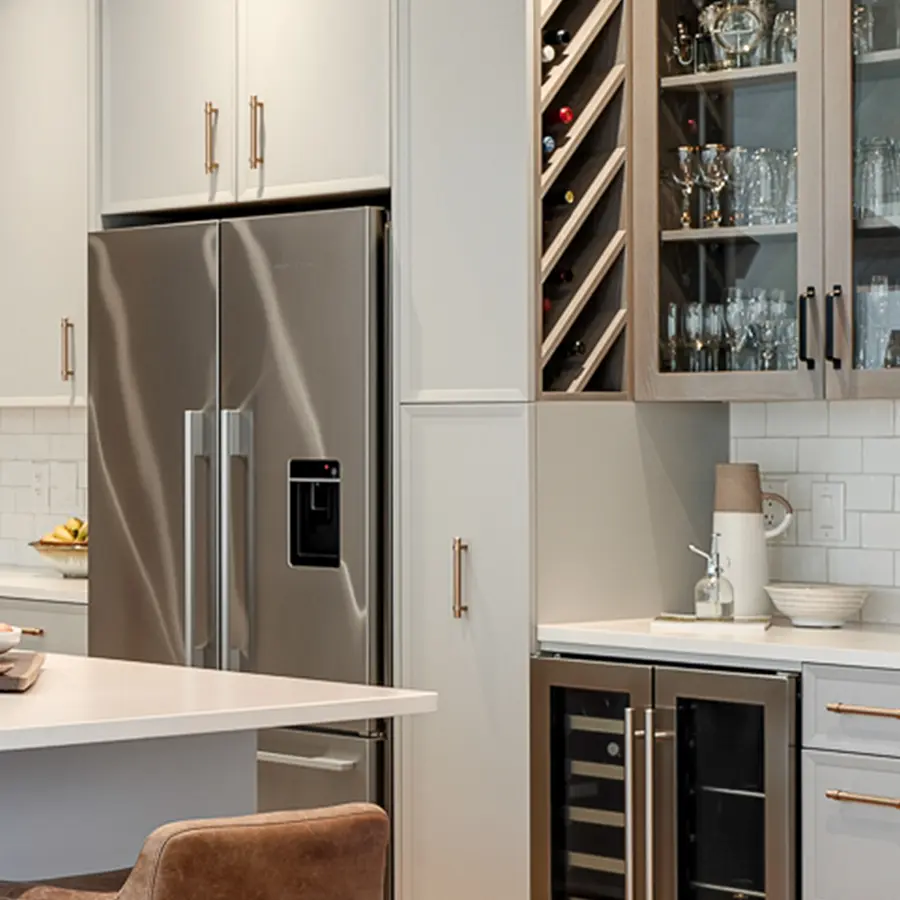 Modern kitchen with stainless steel double-door refrigerator, built-in wine rack, glass-front cabinets displaying glassware, and a wine cooler below the counter. White countertops and subway tile backsplash are visible.