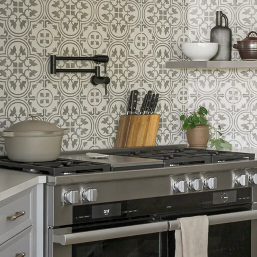A modern kitchen stove with a beige dutch oven, knife block, and potted plant on a counter beneath patterned tile backsplash and open shelves with dishes and bottles. A black pot filler faucet is mounted on the wall.
