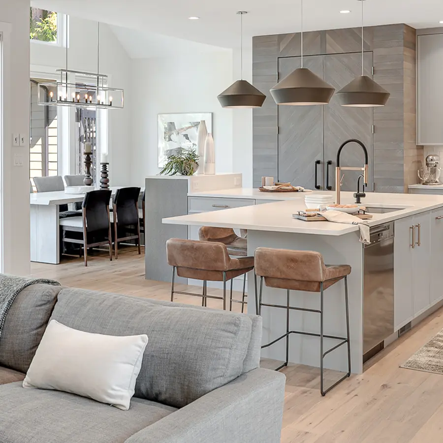 Modern open-concept kitchen and living area with a gray sofa, island with bar stools, pendant lights, light wood flooring, and a dining table in the background. The space features a neutral color palette and contemporary decor.