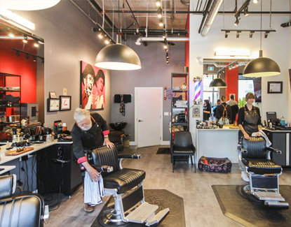 Two barbers stand by black barber chairs in a modern barbershop with red and gray walls, large mirrors, and overhead lights. The shop has hairdressing tools on the counters and framed art on the wall.