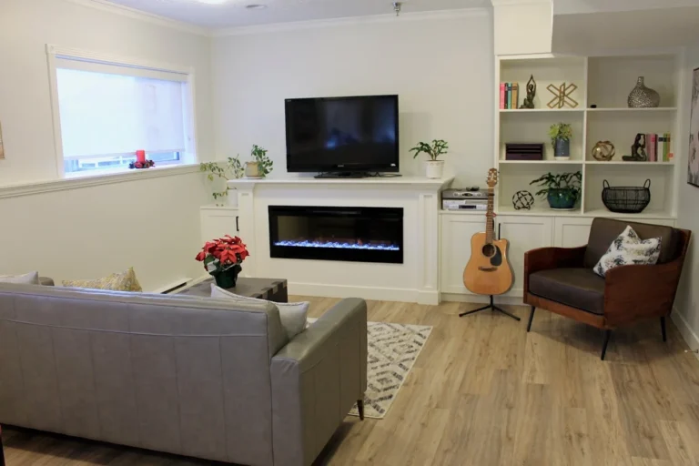 A cozy living room with a gray sofa, brown armchair, guitar, and electric fireplace under a wall-mounted TV. There are shelves with books, plants, and decor on light wood flooring.
