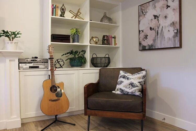 A cozy reading nook with a brown armchair, a patterned pillow, an acoustic guitar on a stand, built-in shelves with books, plants, and decor, and a large floral painting on the wall.