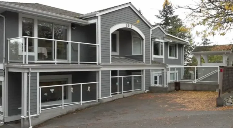 A two-story gray building with white trim and railings, large windows, and a sloped driveway. Some autumn leaves are scattered on the ground, and trees are visible in the background.