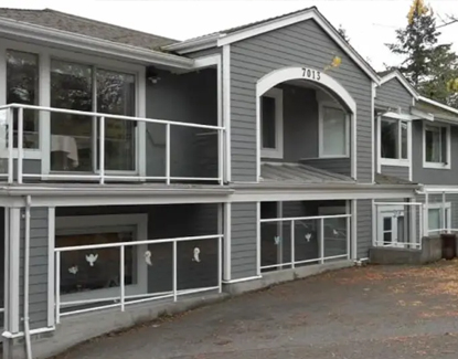 A gray two-story building with white trim and railings, large windows, and the number 7013 above the entrance. The ground and balcony are clear, with a few leaves scattered on the pavement.