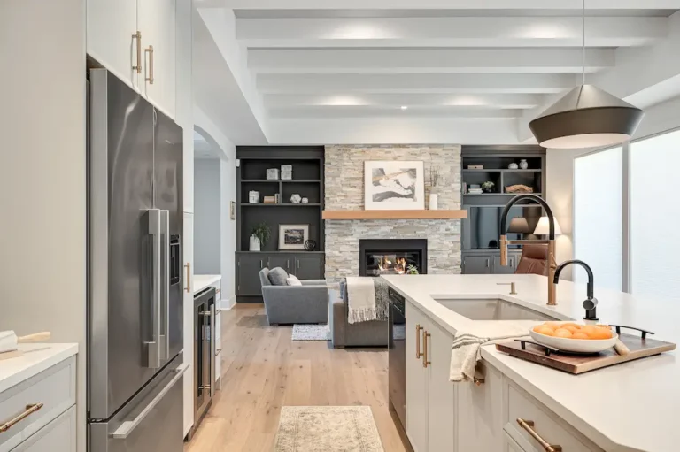 Modern open-concept kitchen and living room with light wood floors, white cabinets, stainless steel appliances, and a stone fireplace. A bowl of oranges sits on the kitchen island, with cozy seating in the background.
