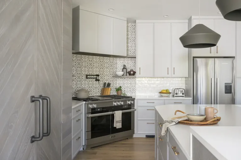 Modern kitchen with white cabinets, patterned backsplash, stainless steel appliances, island with a tray holding dishes, and pendant lights. A refrigerator and toaster are visible next to the countertop.