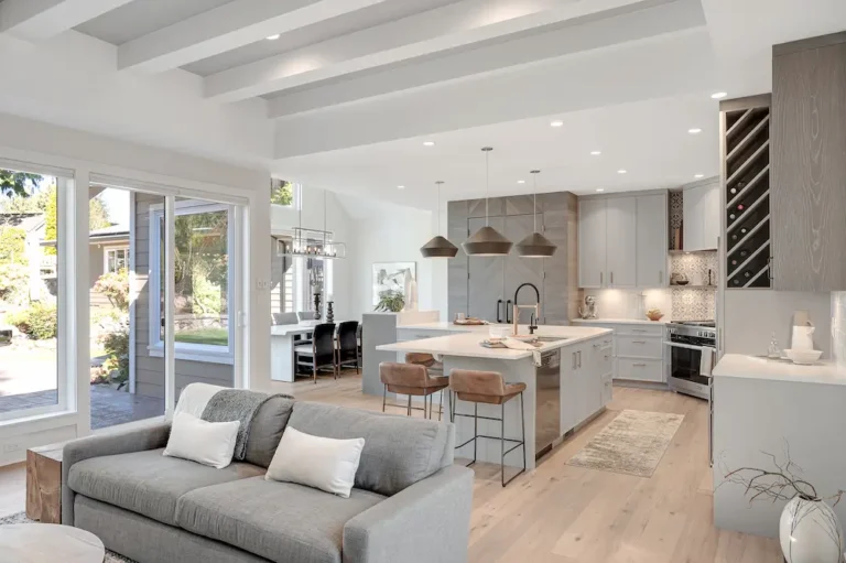 Modern open-concept living room and kitchen with light wood floors, gray sofa, white walls, large island with stools, pendant lights, built-in wine rack, and large sliding doors leading to a sunny backyard patio.
