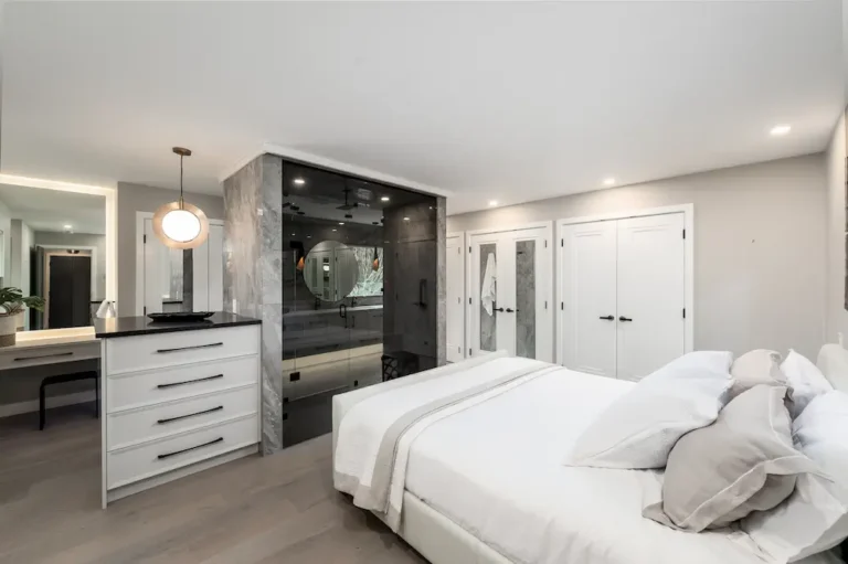 Modern bedroom with a white bed, light grey bedding, a dresser with a round lamp, and a glass-enclosed shower in a marble accent wall. There are white double doors and a vanity with a mirror in the background.