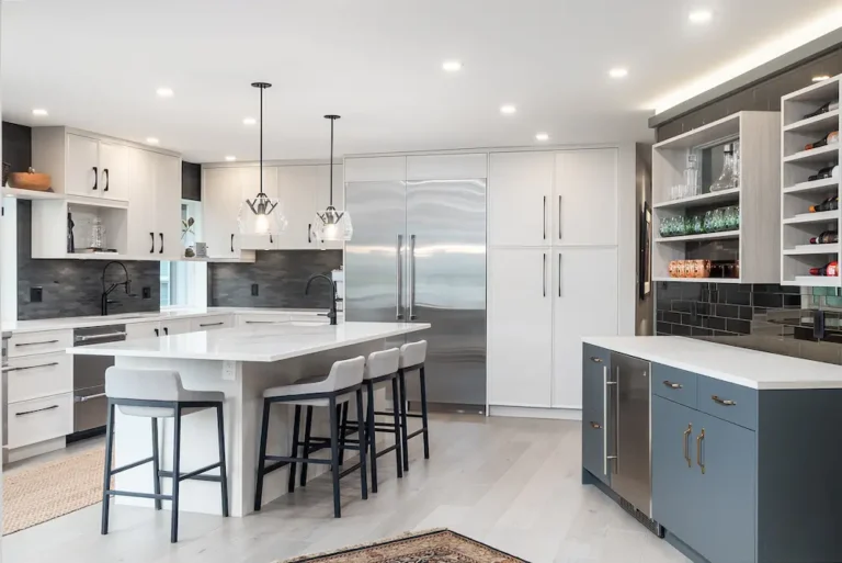 Modern kitchen with white cabinets, stainless steel appliances, a large island with barstools, pendant lights, and open shelves with dishes and glasses. The room features black backsplash and light wood floors.