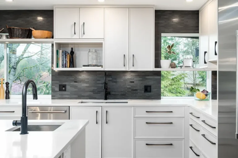 Modern kitchen with white cabinets, black handles, white countertops, and a black hexagonal tile backsplash. Large windows provide a view of green trees outside. The sink and faucet are black, and decor items are on the counters and shelves.