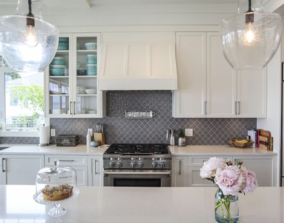 Modern white kitchen with glass-front cabinets, stainless steel stove, gray tile backsplash, pendant lights, a vase with pink flowers, and a cake on a glass stand on the countertop.