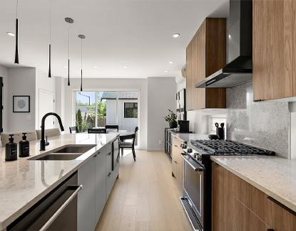 Modern kitchen with a large island, marble countertops, wood cabinets, stainless steel appliances, pendant lights, and a dining area in the background with natural light coming through glass doors.