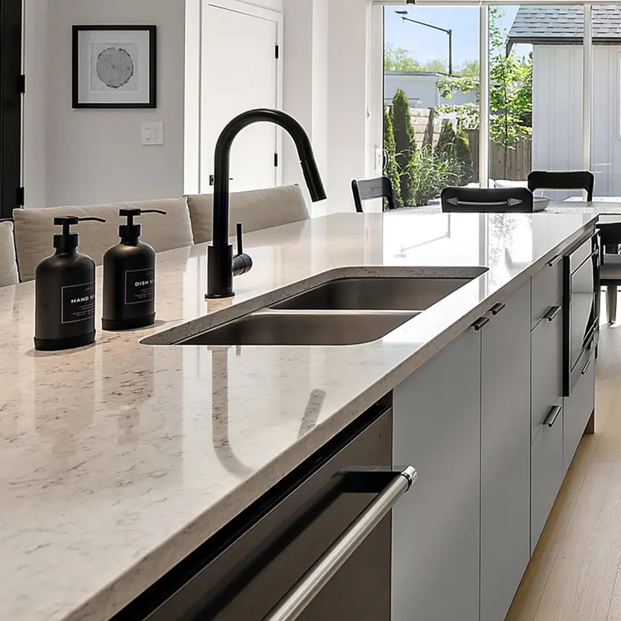 Modern kitchen with a marble island countertop, built-in sink, matte black faucet, and soap dispensers. Neutral cabinets and bar stools line the island, and large windows provide natural light and a view of a fenced backyard.