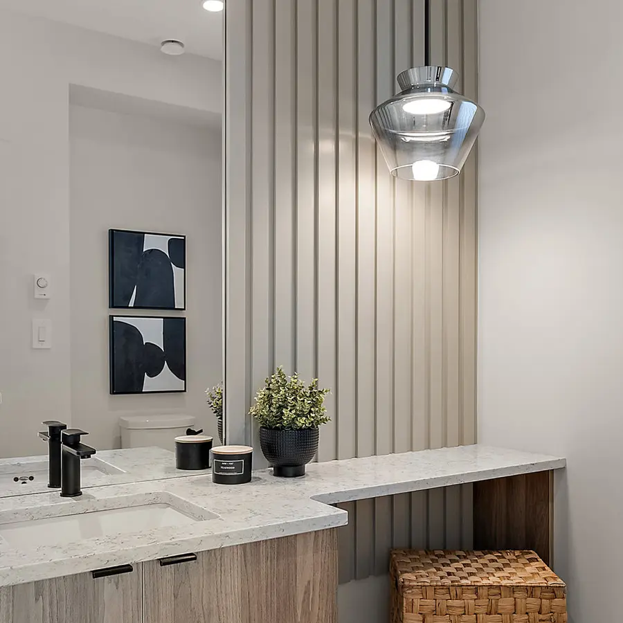 A modern bathroom with a marble countertop, black fixtures, a potted plant, wicker basket, and a hanging glass light. Vertical slats and abstract art decorate the neutral-toned space.