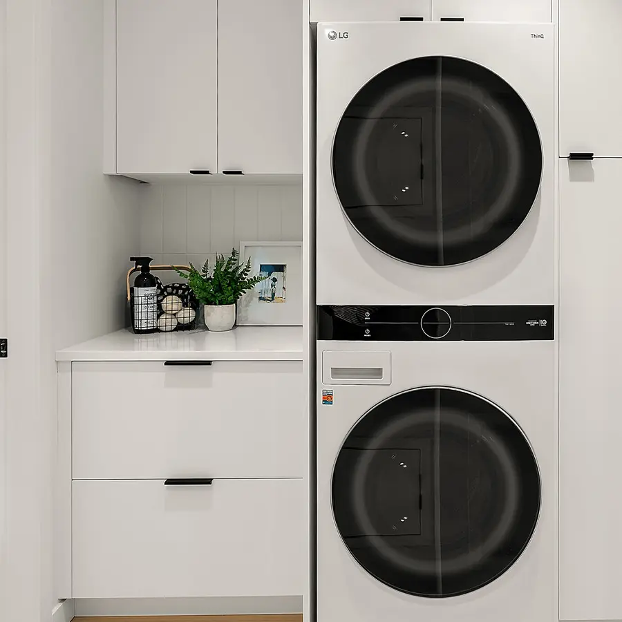 A modern laundry area with a stacked LG washer and dryer, white cabinets, drawers with black handles, a countertop with a potted plant, framed photo, soap pump, and a wire basket with laundry accessories.