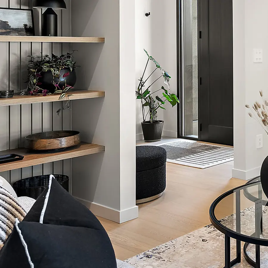 Modern living room with light wood floors, black and neutral decor, open shelving with plants and bowls, round glass coffee table, a black ottoman, and a potted plant near a doorway with natural light.