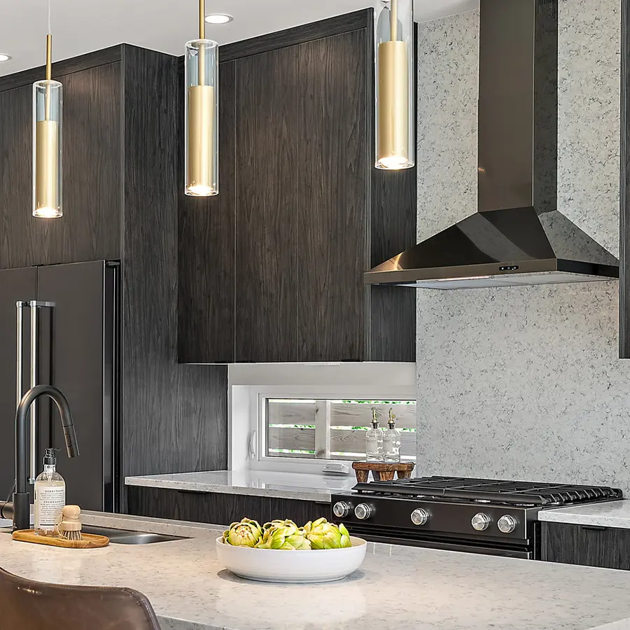 Modern kitchen with dark wood cabinets, a black refrigerator, and a black range hood over a stove. Three gold pendant lights hang above a marble island with a sink and a bowl of green artichokes as decor.
