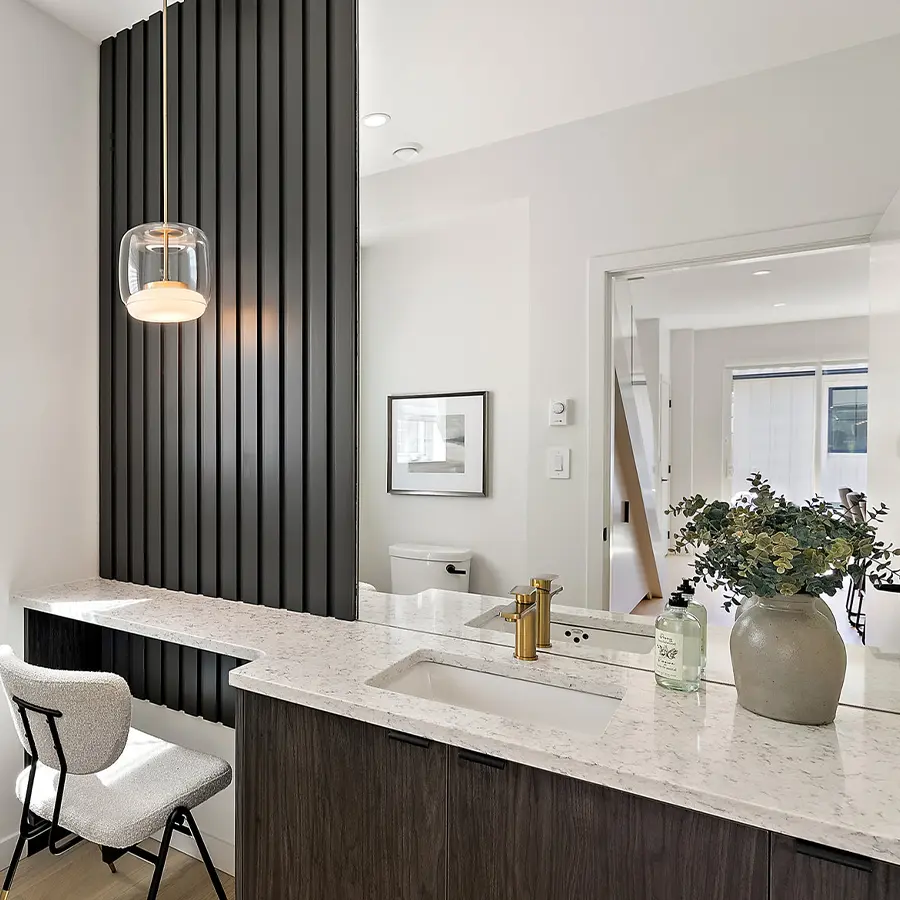 Modern bathroom with a marble countertop, gold faucet, large mirror, decorative vase with greenery, black vertical wall slats, a white chair, pendant light, and neutral-colored walls.