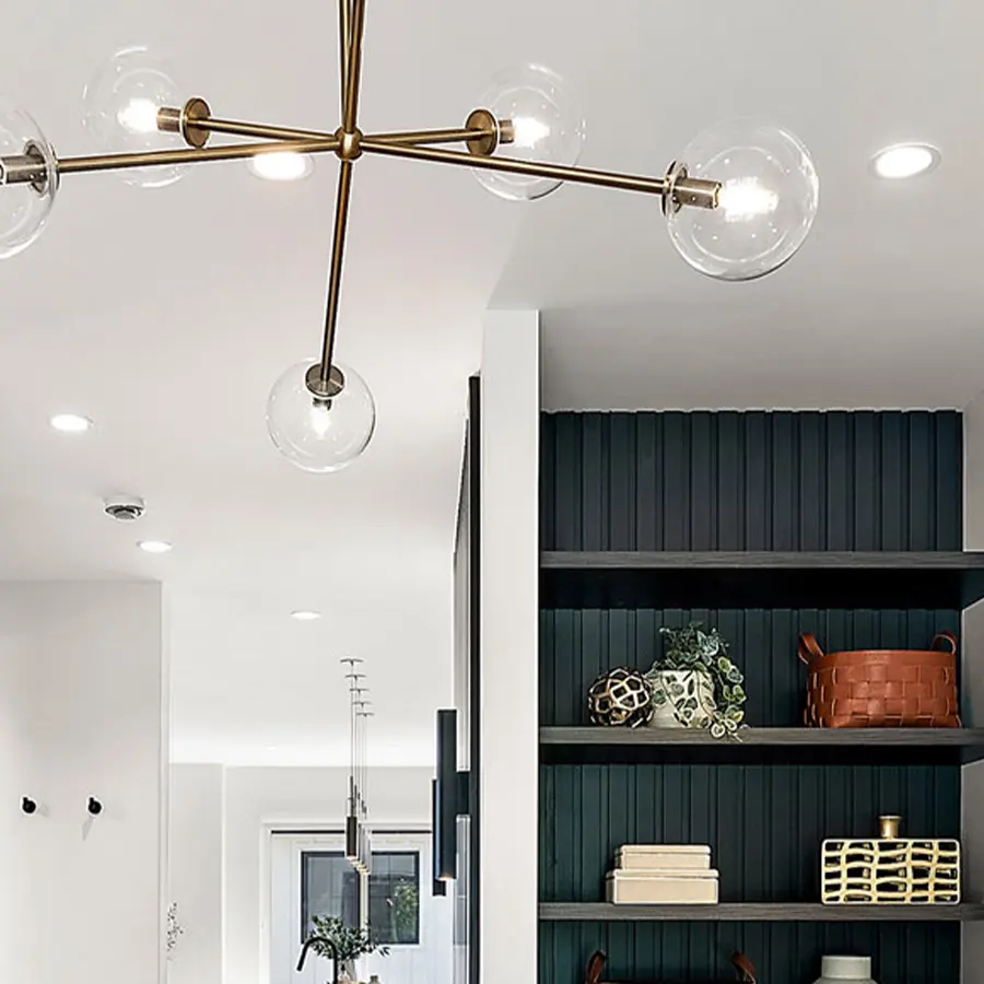 A modern interior with a brass chandelier featuring exposed bulbs, built-in shelves with decor items, and a white wall. The shelves hold books, plants, vases, and a woven basket.