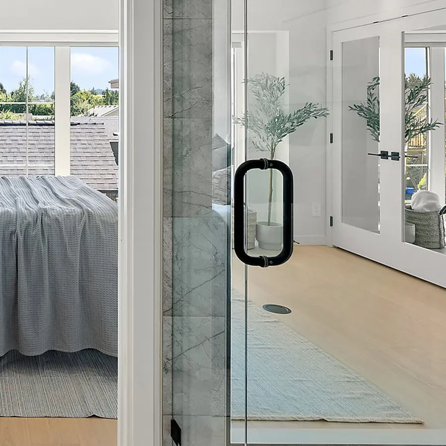 A modern bedroom with glass doors, light wood floors, a neatly made bed with a gray textured blanket, large windows with a view of greenery, and a potted plant in the corner.
