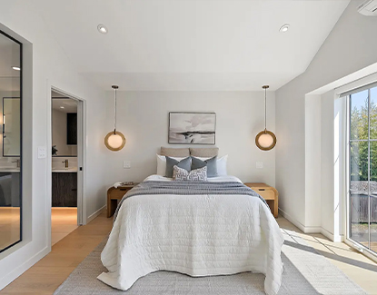 A bright, modern bedroom with a neatly made bed, white bedding, and two pendant lights hanging above wooden nightstands. Large windows let in natural light, and a bathroom is visible through an open door on the left.