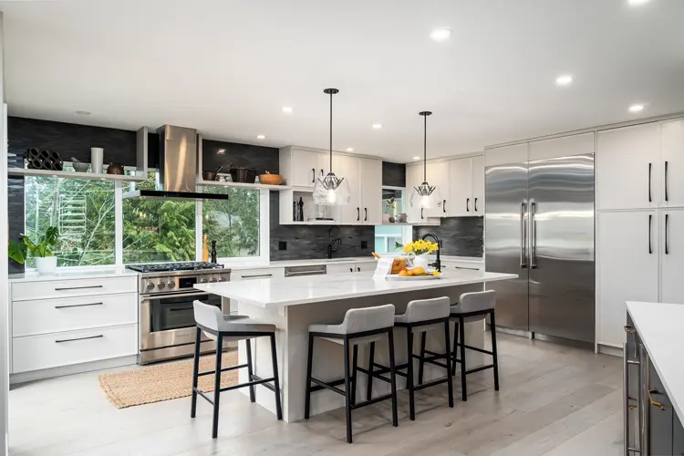 Modern kitchen with white cabinets, stainless steel appliances, a large island with four gray stools, pendant lights, and a window over the sink showing greenery outside. Countertops are white, and the floor is light wood.
