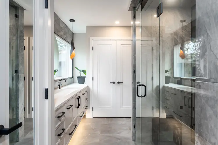 Modern bathroom with gray marble floors and walls, glass shower enclosure, white cabinets with black handles, under-cabinet lighting, a rectangular sink, and a large window with a hanging pendant light.