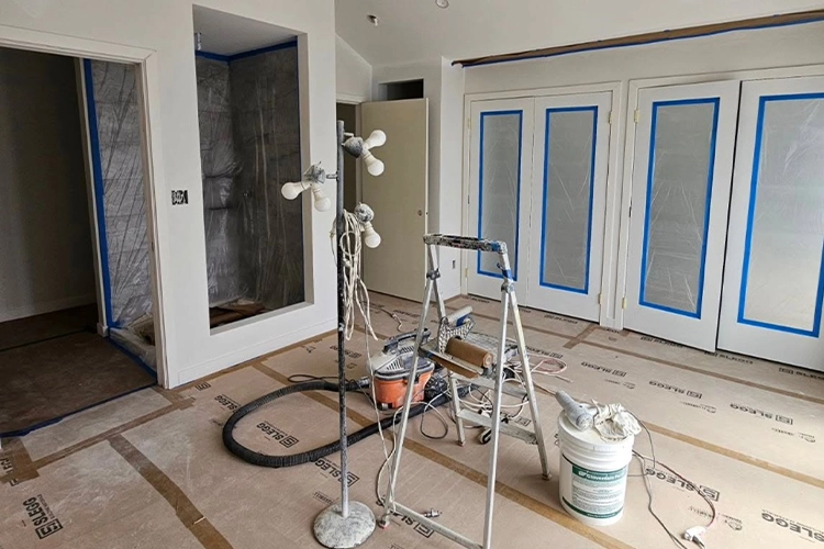 A room under renovation with floors covered in protective sheets, a ladder, painters tape on doors and windows, work lights, a bucket, and painting equipment scattered on the floor.