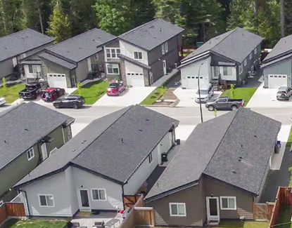 Aerial view of a suburban neighborhood with modern, single-family homes, driveways, and cars parked outside. The area is surrounded by trees and greenery.