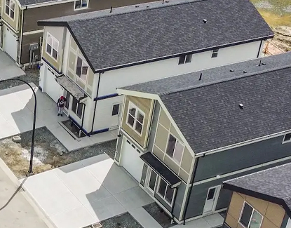 Aerial view of two modern, two-story townhouses with dark roofs and attached garages. A person stands near the entrance of one house. The area around the houses is paved with driveways and sidewalks.