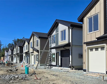 A row of newly constructed two-story houses with garages, ladders, and construction materials visible, set against a clear blue sky. The ground is unfinished, indicating ongoing construction.