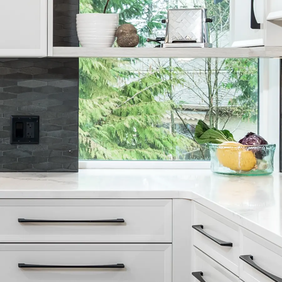 A modern white kitchen with black handles, a grey textured backsplash, and a bowl of produce by a large window showing green trees outside. White dishes and decor sit on an upper shelf.