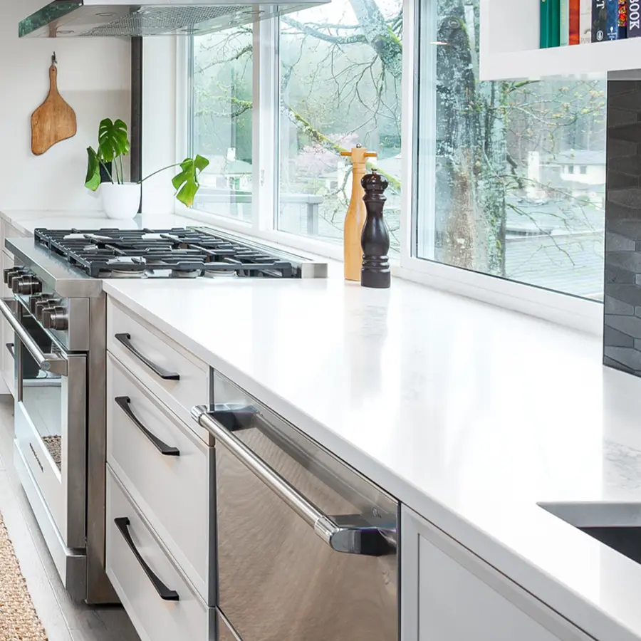A modern kitchen with white countertops, a stainless steel gas stove, dishwasher, and large windows. A potted plant, cutting board, and salt and pepper grinders are on the counter. Trees are visible outside the window.