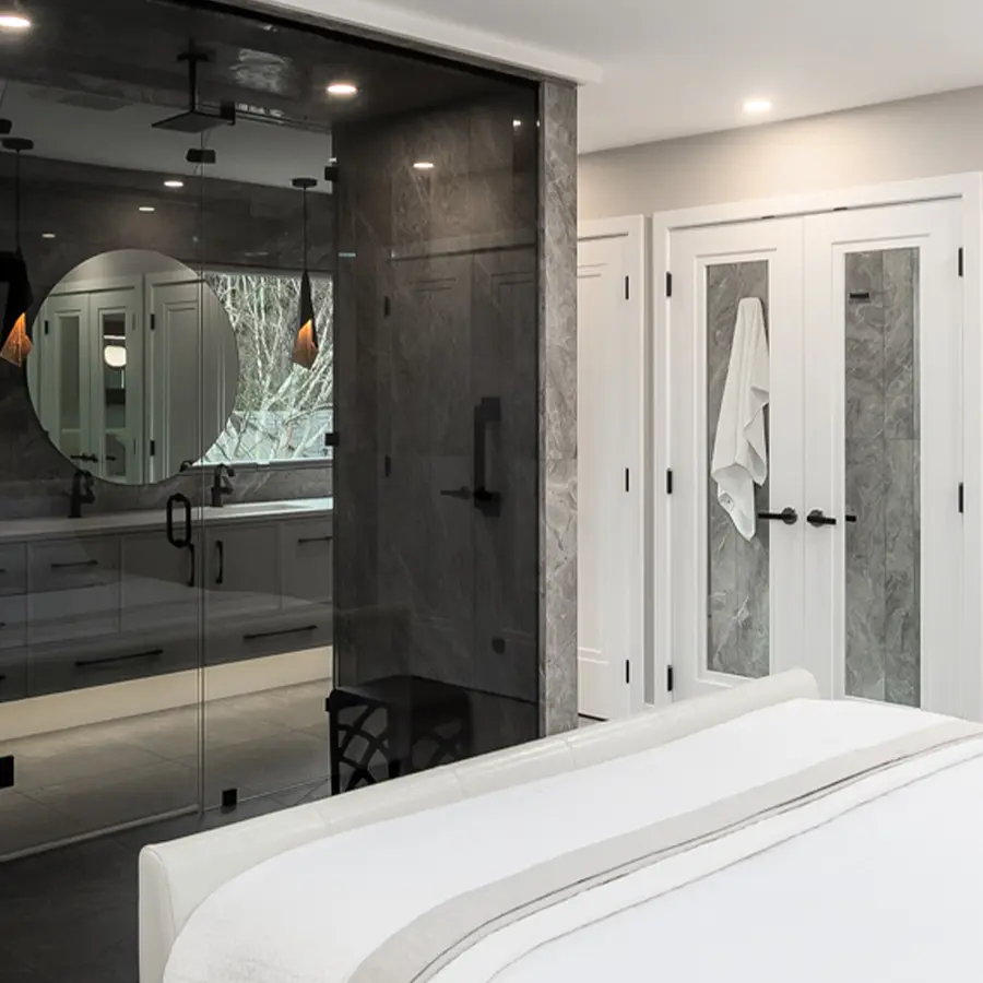 Modern bedroom with a view into a glass-walled bathroom featuring a round mirror, gray tiles, and a walk-in shower. White double doors with a hanging towel are at the far end of the room.