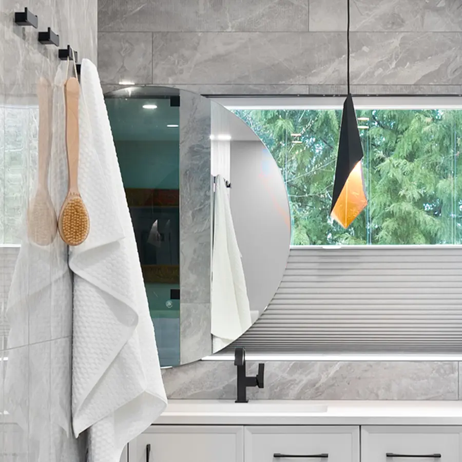 Modern bathroom with gray marble walls, a round mirror, and a window with a view of green trees. A pendant light hangs above a white counter, and towels with wooden brushes are hanging on hooks on the left.