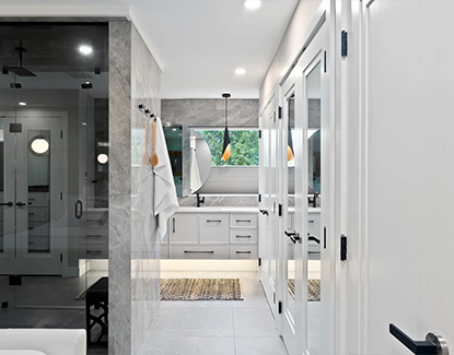 Modern bathroom with gray tile floors, white cabinetry, a large mirror, pendant lights, a glass-enclosed shower, and towels hanging on the wall. Natural light enters through a window above the sink.